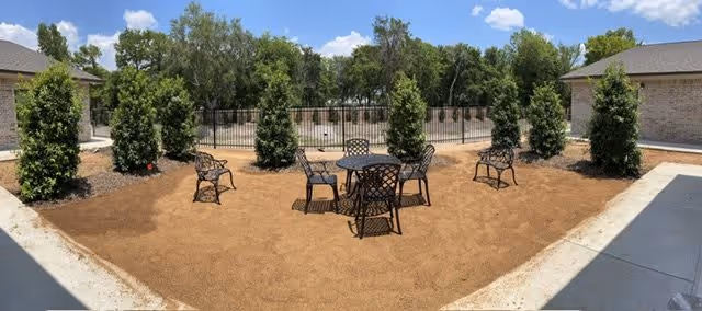 Outdoor patio area with a round metal table and four matching chairs on a dirt ground, surrounded by small green bushes and trees in the background under a blue sky with some clouds. Two buildings with brick walls and gray roofs are visible on either side of the patio.