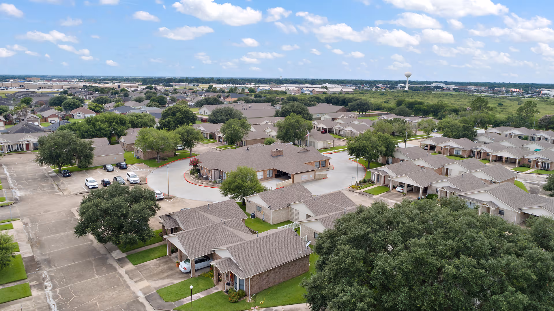 Aerial view of a suburban senior living community with single-story homes, driveways, trees, and parking areas under a partly cloudy sky.