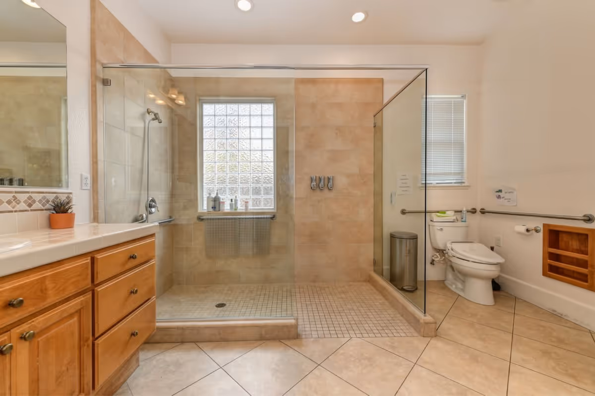 A spacious bathroom with a large walk-in glass shower featuring beige tiles and a window with frosted glass blocks. To the right is a toilet with grab bars on the walls, a small window with blinds, a trash can, and a built-in wooden shelf. On the left side, there is a wooden vanity with drawers and a countertop with a small potted plant.
