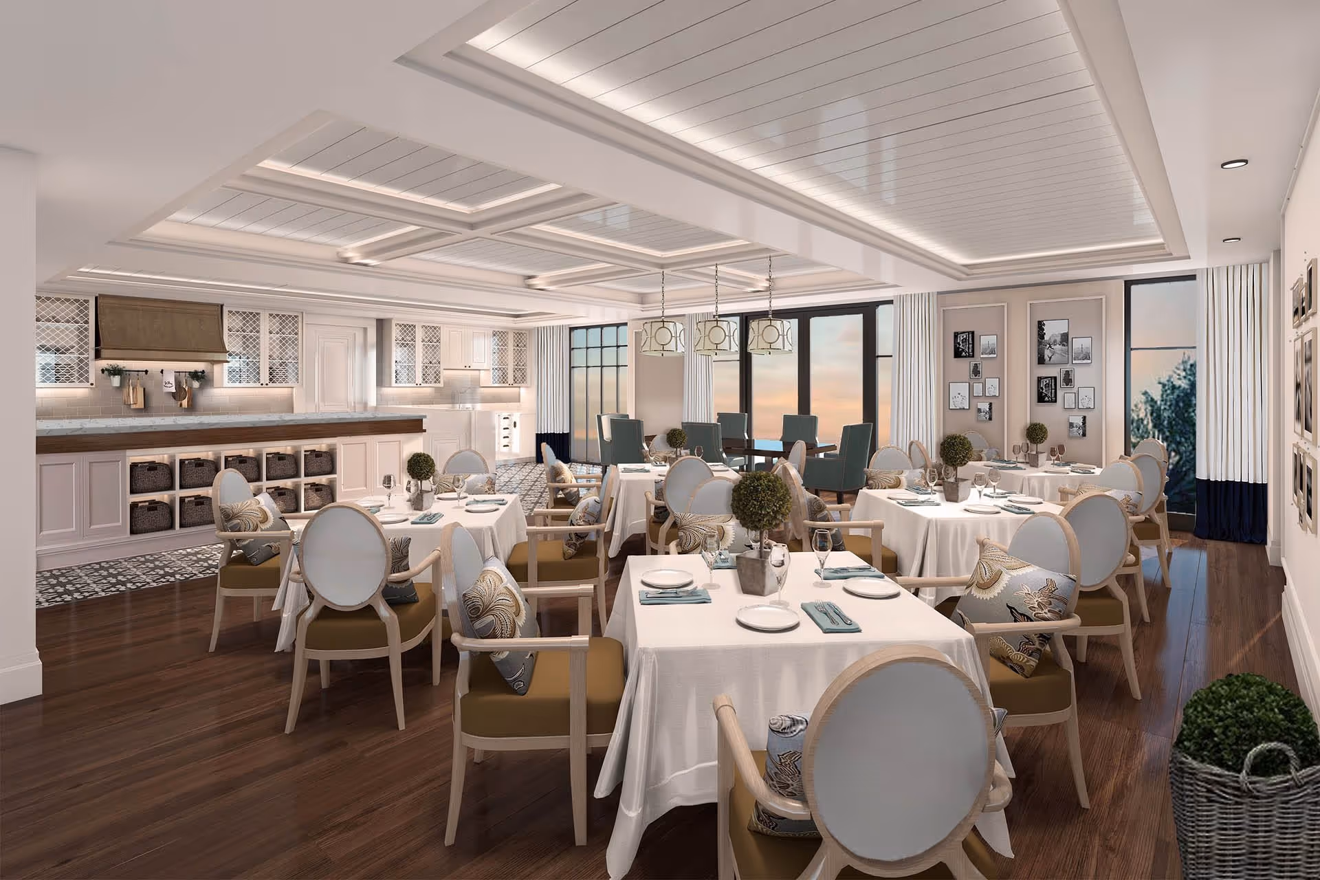 A bright and elegant dining room with multiple tables covered in white tablecloths, each set with plates, glasses, and napkins. The chairs have light wooden frames with cushioned seats and decorative pillows. The room features large windows with white and navy curtains, a coffered ceiling with recessed lighting, and a kitchen area in the background with white cabinetry and a wooden countertop. The floor is dark wood, and there are small decorative plants on the tables and in a basket on the floor.