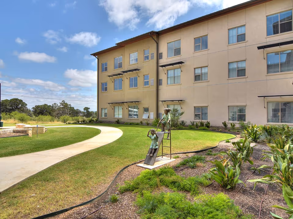 Outdoor view of a senior living facility building with a curved concrete walkway, green lawn, landscaped garden with plants, and a bronze sculpture of two children playing on a slide under a partly cloudy blue sky.