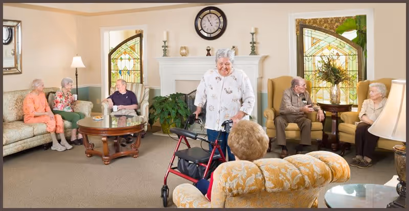 A group of elderly residents socializing in a bright common living room with upholstered chairs, a fireplace, and stained-glass windows.