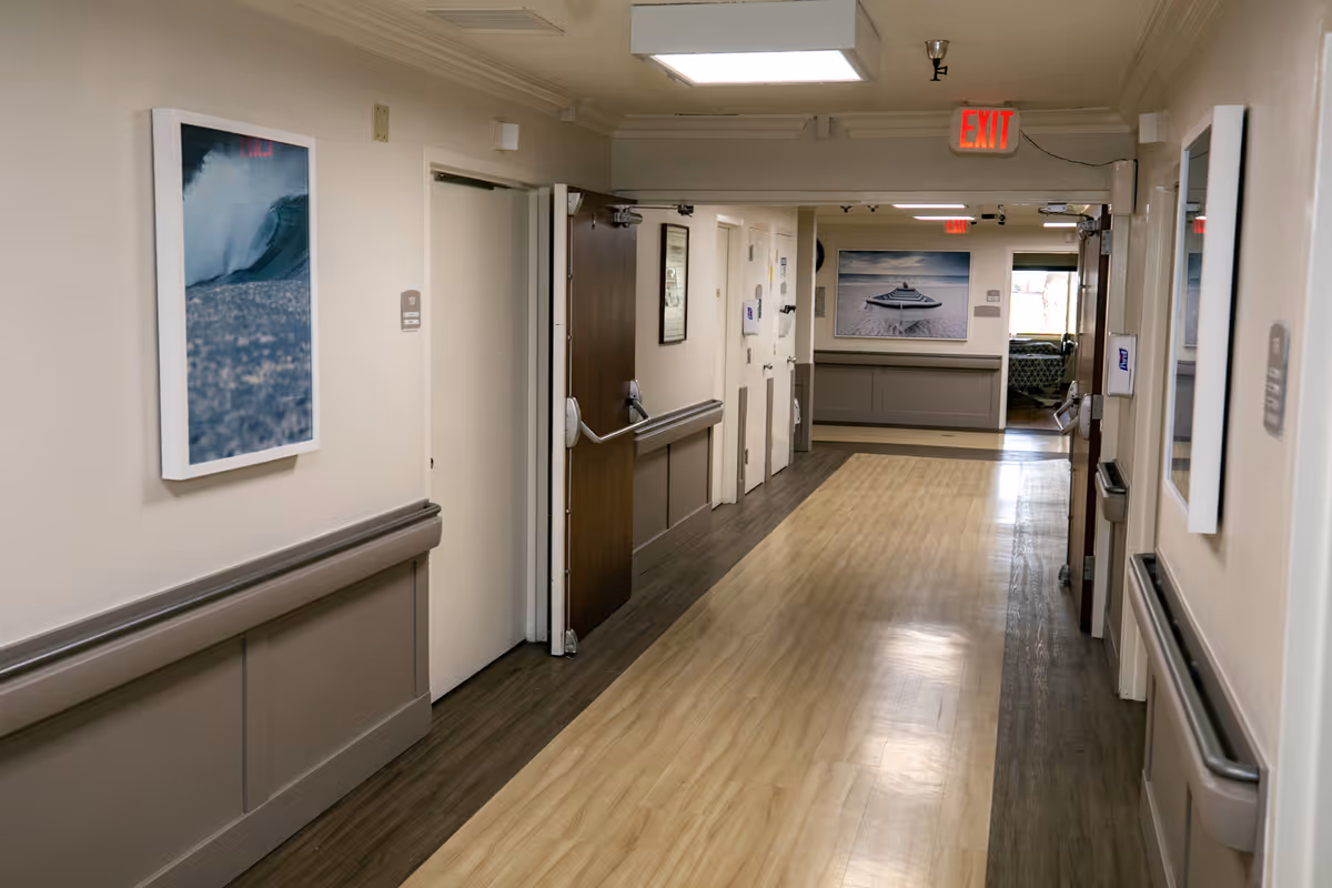 Well-lit interior hallway in a healthcare facility with handrails, doors, framed artwork, and an exit sign.