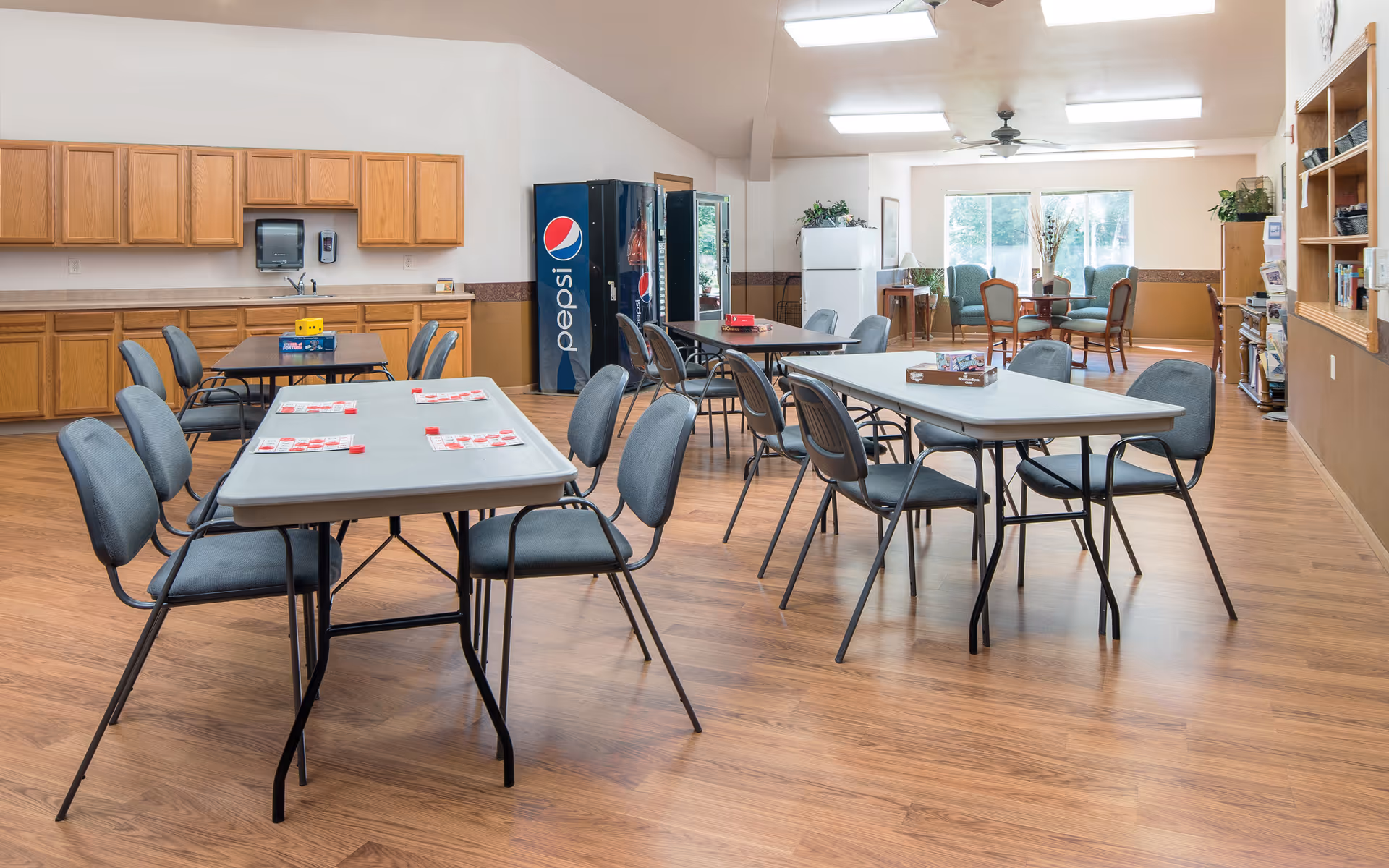 A spacious common area with multiple tables and chairs arranged for group activities or dining. The room has wooden flooring, a Pepsi vending machine, a refrigerator, and cabinets along one wall. In the background, there is a seating area with armchairs and a round table near large windows letting in natural light.