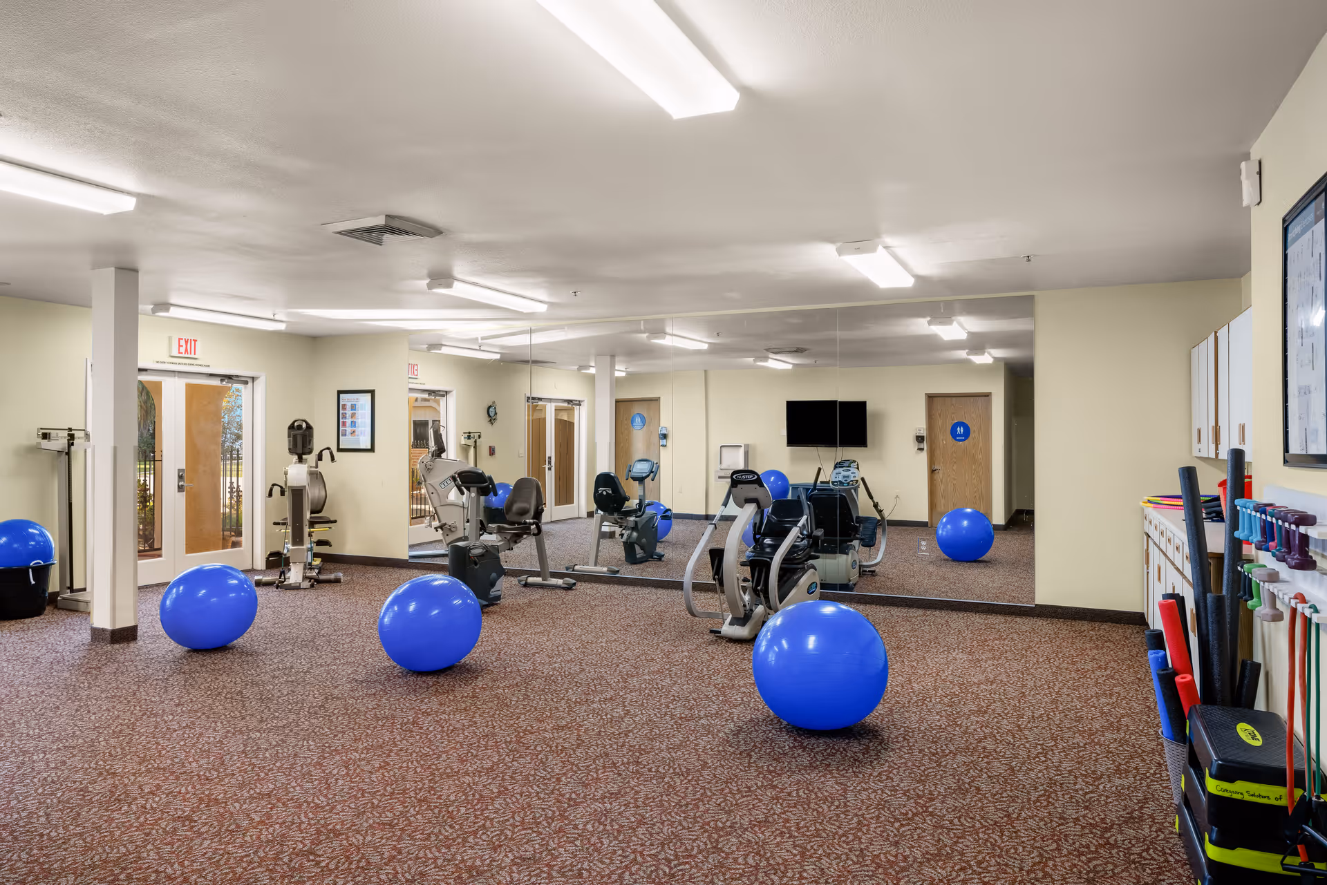 A senior living community exercise room with several blue exercise balls, stationary exercise bikes, a large wall mirror, and various exercise equipment organized on shelves. The room has beige walls, carpeted floor, and fluorescent ceiling lights. There are doors and an exit sign visible in the background.