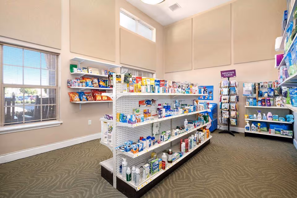 Interior view of a small convenience or sundries store inside a facility, with shelves stocked with various personal care products, snacks, and greeting cards. The room has large windows with blinds, beige walls, and carpeted flooring.
