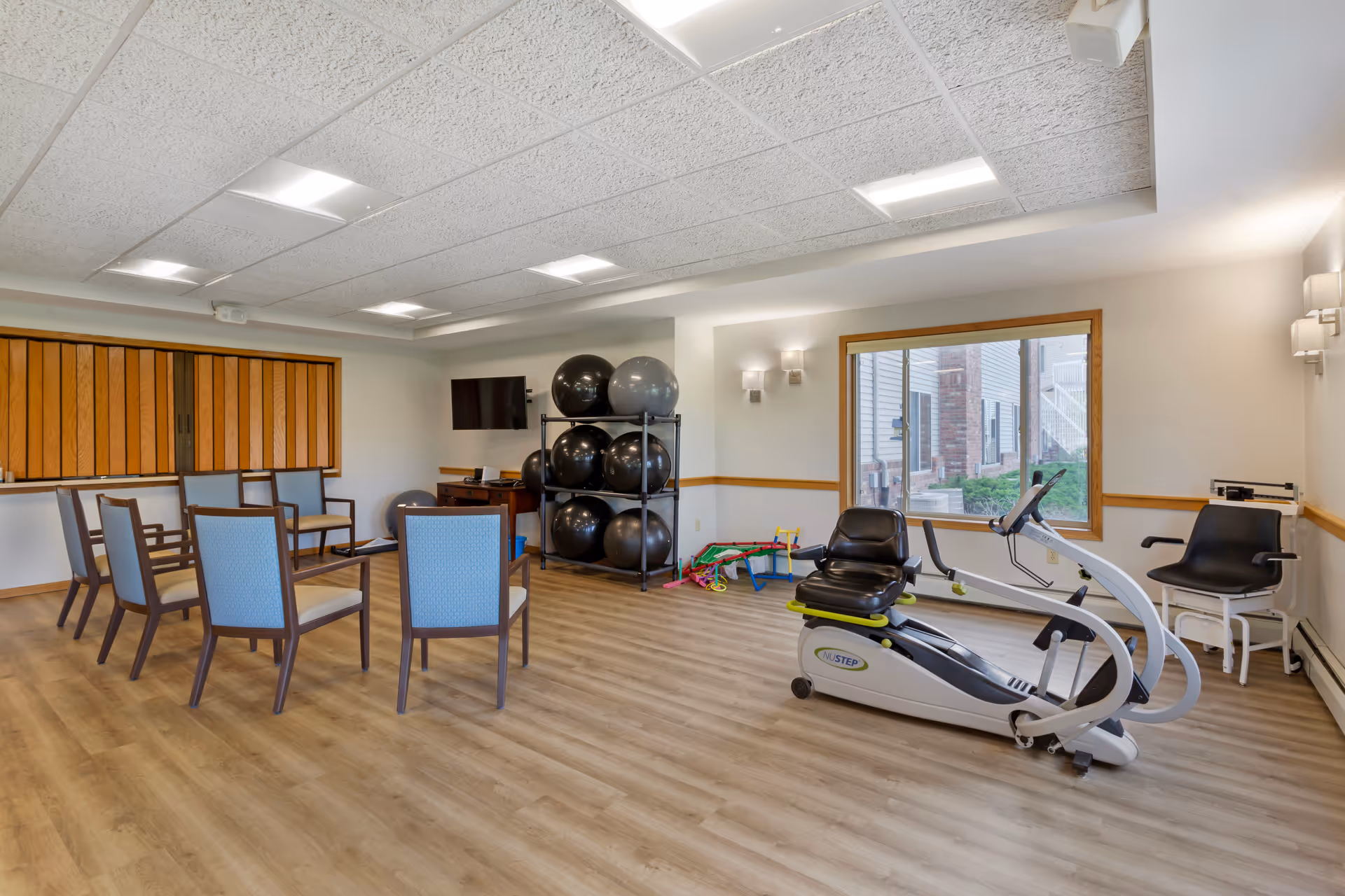 A senior living facility exercise room with wooden flooring, several chairs arranged in a semi-circle, a rack holding large exercise balls, a recumbent exercise bike, and a window showing an exterior view of the building.