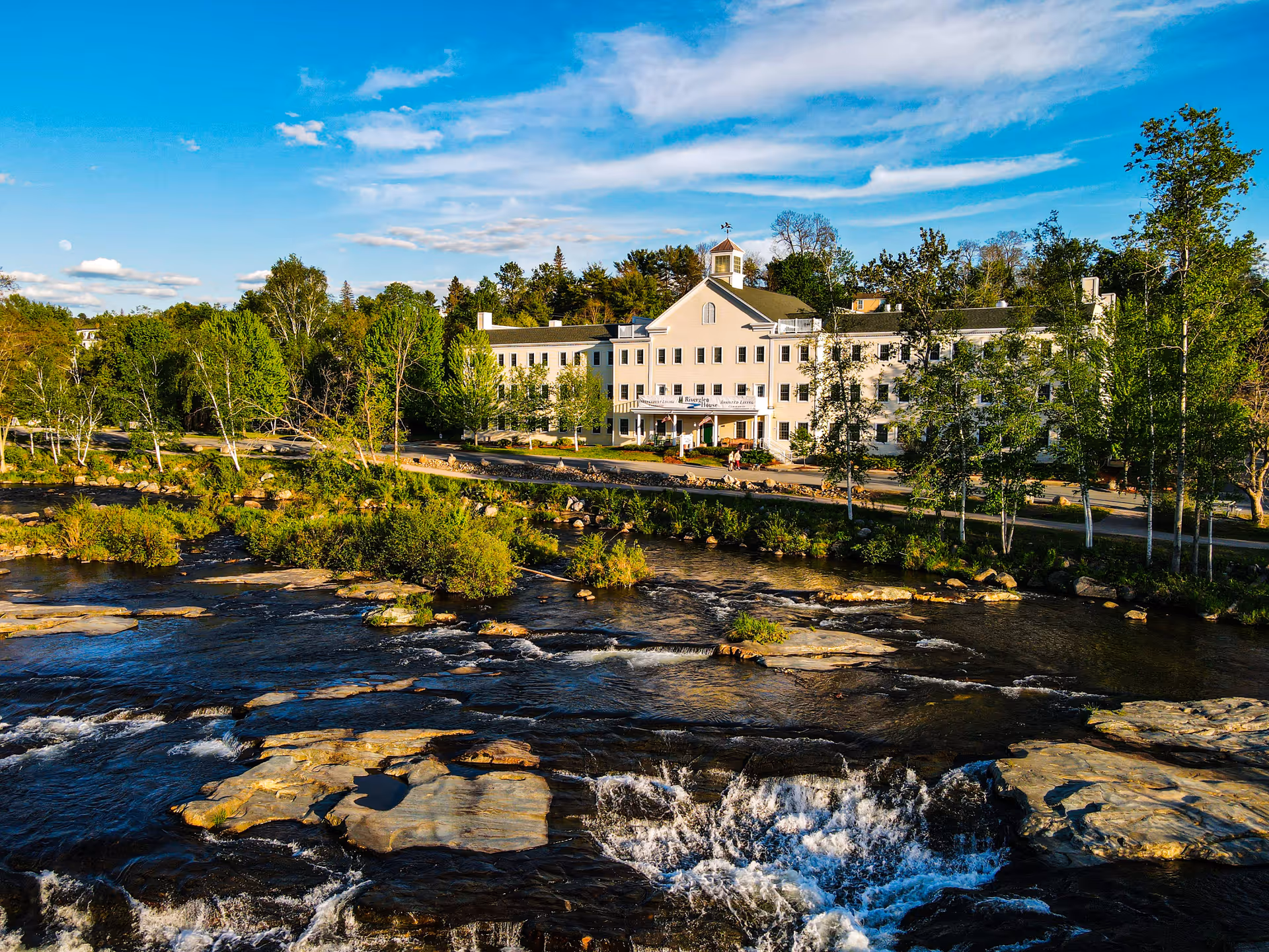 A large, multi-story white building named Riverglen House situated beside a flowing river with rocks and small rapids. The building is surrounded by green trees under a blue sky with scattered clouds.
