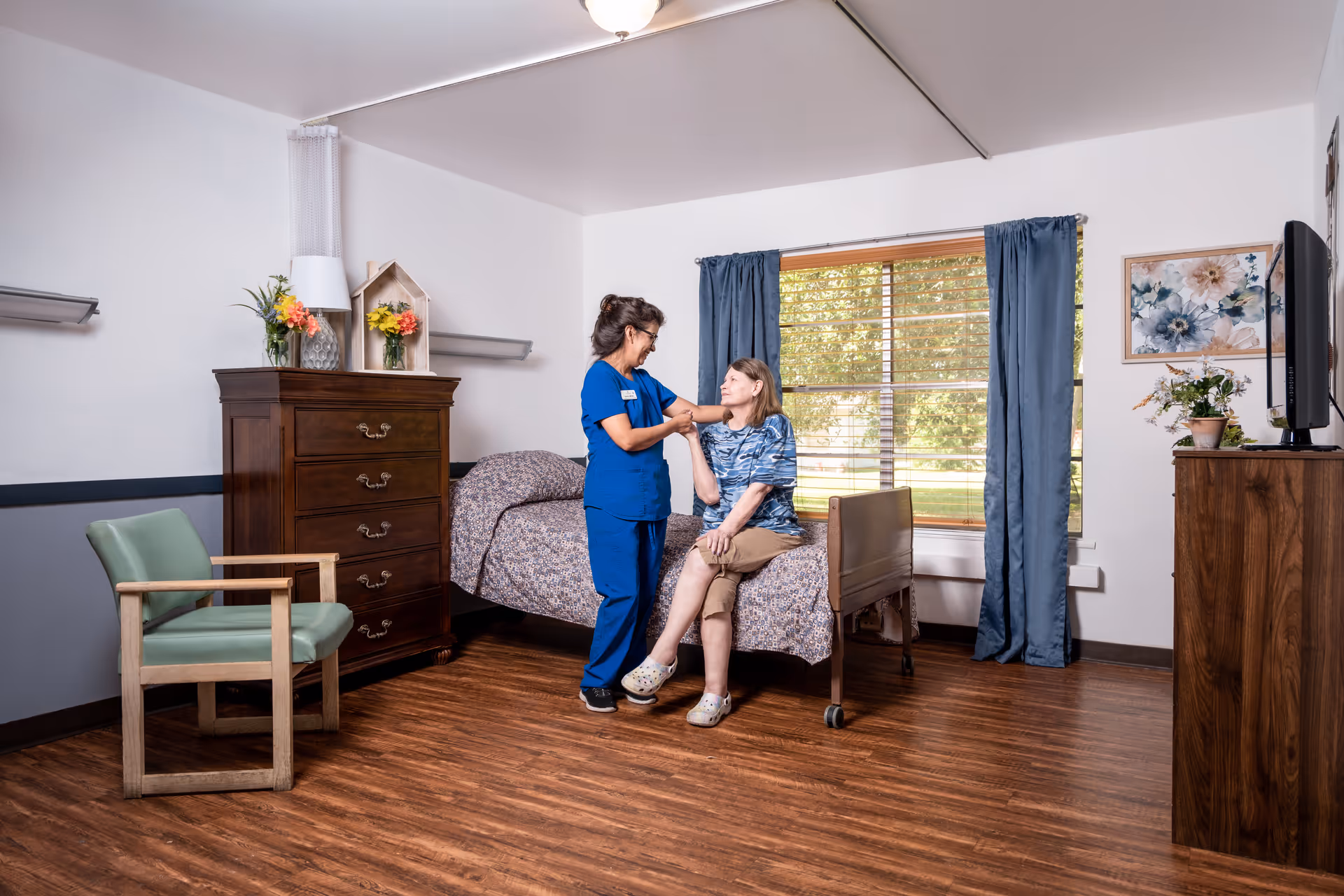 A nurse in blue scrubs is assisting an elderly woman sitting on a bed in a well-lit room with wooden flooring. The room has a wooden dresser with flowers and a lamp, a green cushioned chair, a window with blue curtains, and a TV on a wooden stand. The elderly woman is wearing a blue patterned shirt and beige pants.