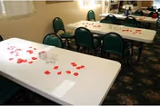 Interior view of a dining area with white rectangular tables and green chairs. The tables have scattered red rose petals and small glass candle holders. The room has carpeted flooring and light-colored walls with windows.