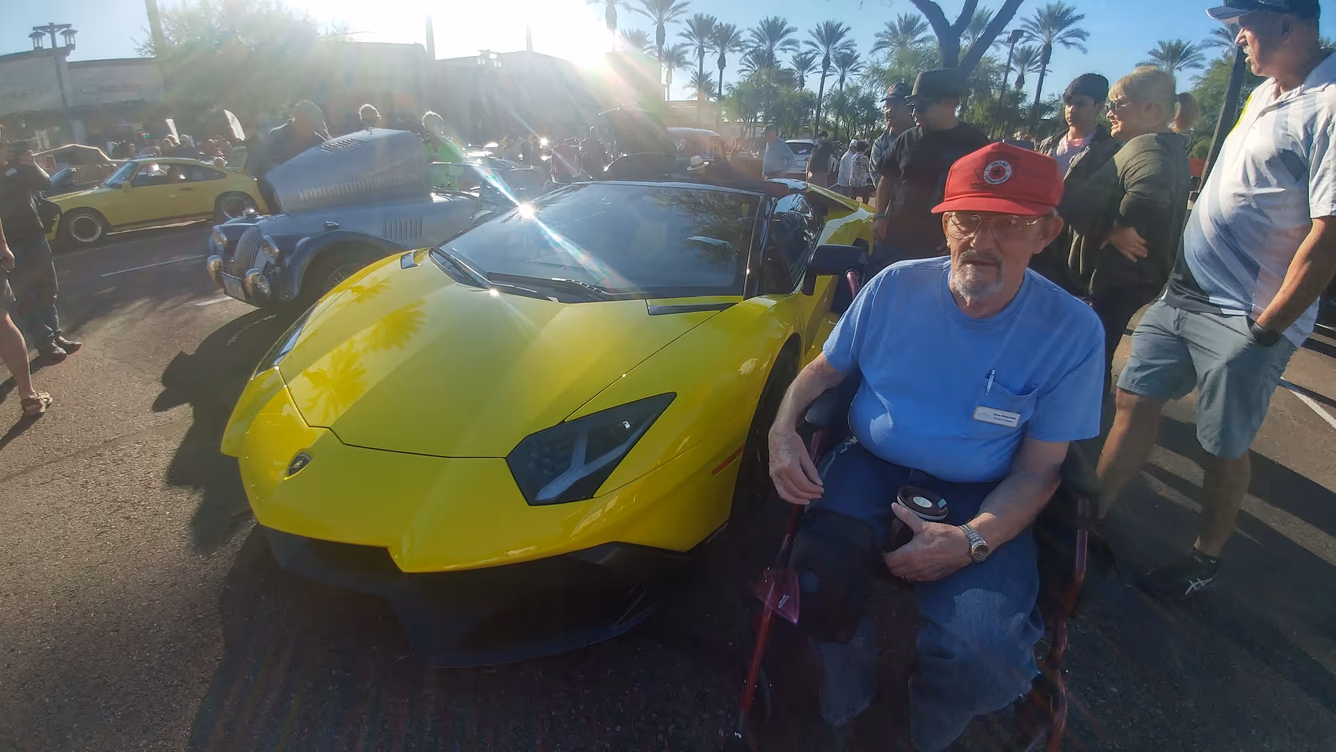 An older man in a wheelchair holding a cup sits beside a yellow Lamborghini at an outdoor car show with palm trees and a crowd in the background.