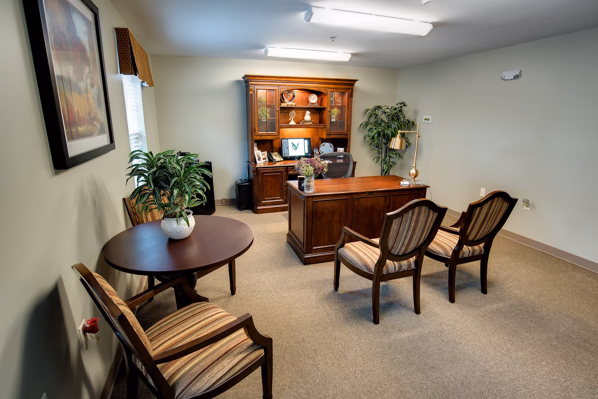An office room with a wooden desk and hutch, two striped upholstered chairs facing the desk, a round table with two matching chairs, a potted plant on the table, a framed painting on the wall, and a large potted plant in the corner.