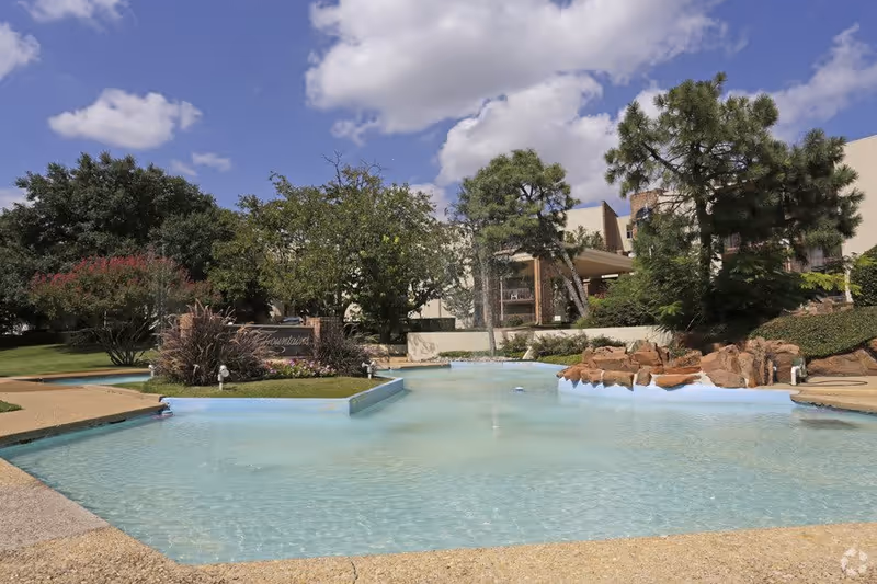 Outdoor view of a senior living facility named Three Fountains featuring a large decorative water pool with clear blue water, surrounded by trees, bushes, and landscaped greenery under a partly cloudy sky.