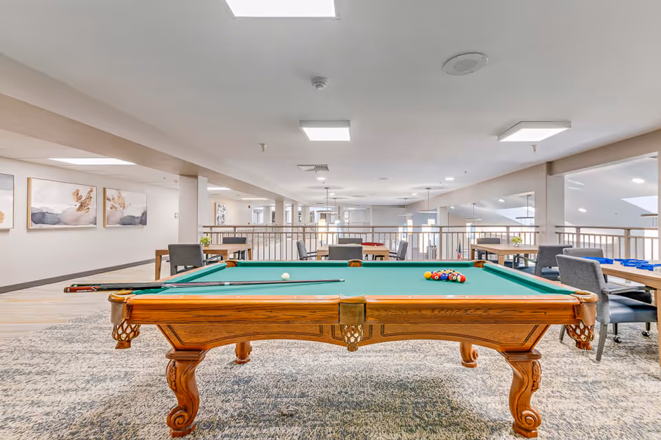 A spacious indoor recreational area featuring a wooden pool table with green felt, pool cues, and a rack of billiard balls set up for a game. Surrounding the pool table are several tables and chairs, with modern lighting and artwork on the walls in the background. The area has a light and airy atmosphere with a carpeted floor and railings overlooking a lower level.
