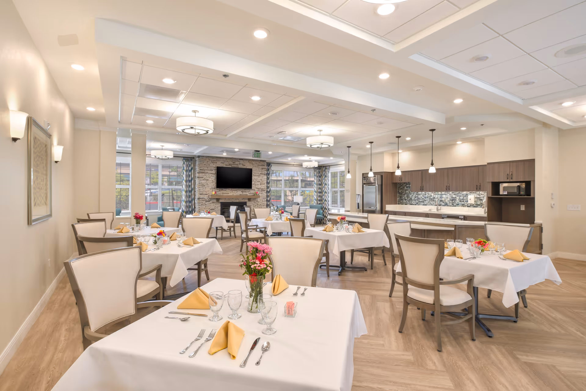 A bright and spacious dining room in a senior living facility with multiple tables covered in white tablecloths, each set with yellow napkins, glassware, and silverware. The room features comfortable chairs, a modern kitchen area with pendant lights, a stone fireplace with a mounted TV, and large windows with patterned curtains allowing natural light to fill the space.