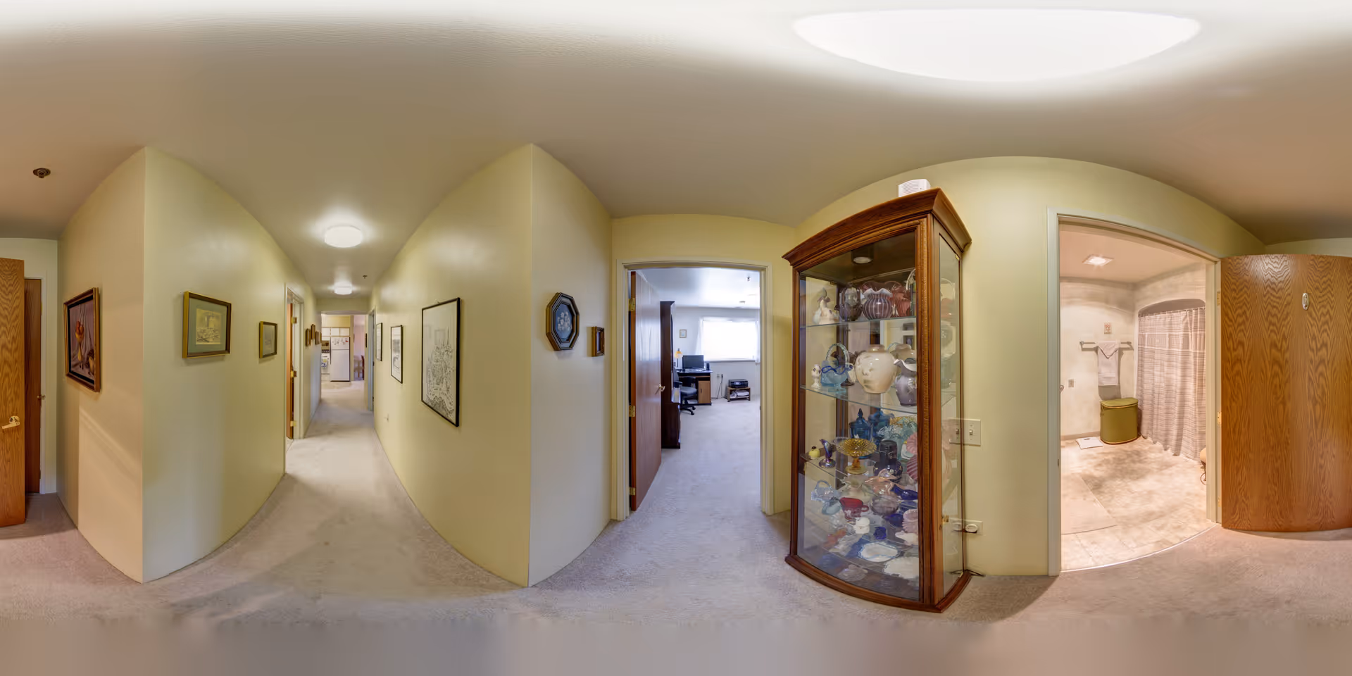 Panoramic interior hallway with carpeted floor, framed artwork on the walls, a glass display cabinet and open doorways including a bathroom.