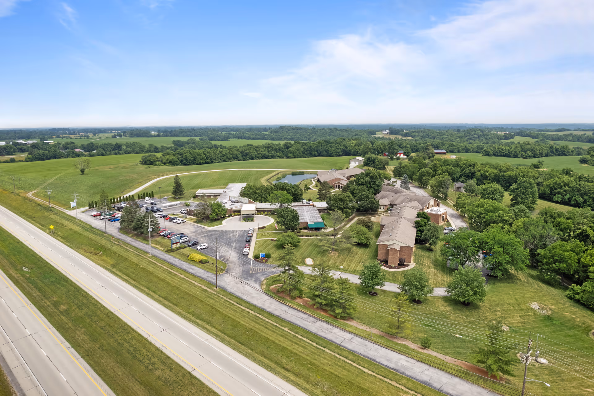 Aerial view of a nursing and rehabilitation campus next to a highway with parking lots, landscaped grounds, and surrounding green fields.