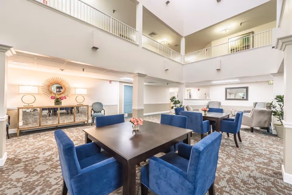 Bright and spacious common area in a senior living facility with high ceilings and an open second-floor balcony. The room features dark wooden tables surrounded by blue upholstered chairs, beige patterned carpet, and decorative elements including a sunburst mirror, table lamps, and small flower arrangements on the tables. There are additional seating areas with armchairs and plants, creating a welcoming and comfortable atmosphere.