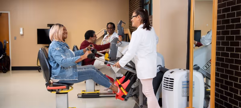 A woman in a denim jacket exercises on a seated leg press machine while a healthcare worker in a white coat assists her. Two other women are seated on exercise machines in the background in a rehabilitation facility room.