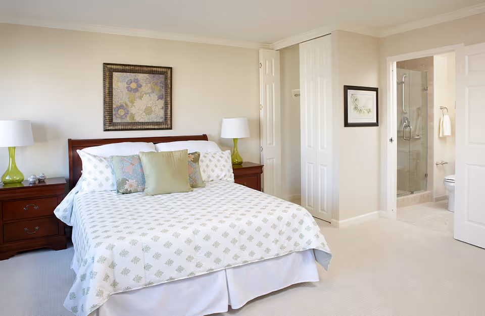 A bright bedroom with a large bed covered in white and green patterned bedding, two green glass lamps on wooden nightstands on either side of the bed, a framed floral artwork above the headboard, an open closet with white bi-fold doors, and an open door leading to a bathroom with a glass shower enclosure and a toilet.