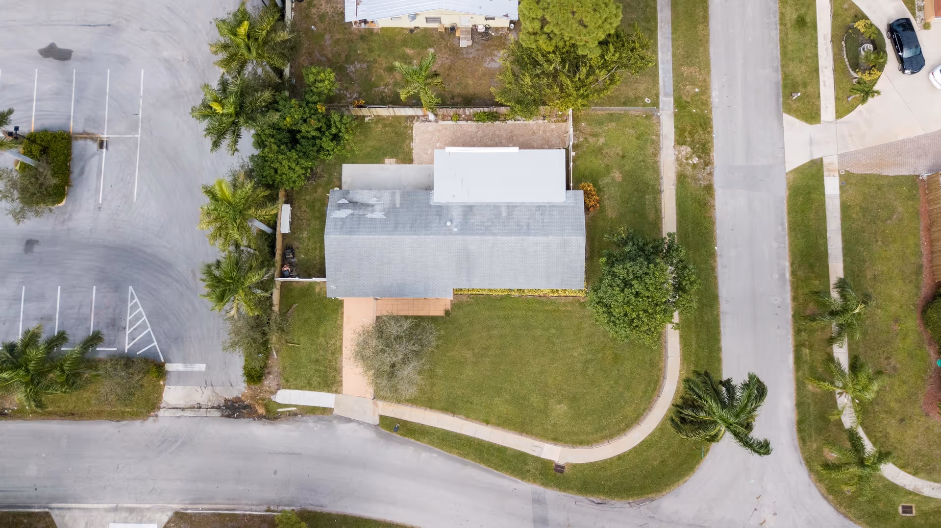 Aerial view of a single-story building with a gray roof surrounded by green lawns, palm trees, and paved roads. There is a parking lot with marked spaces on the left side of the building.