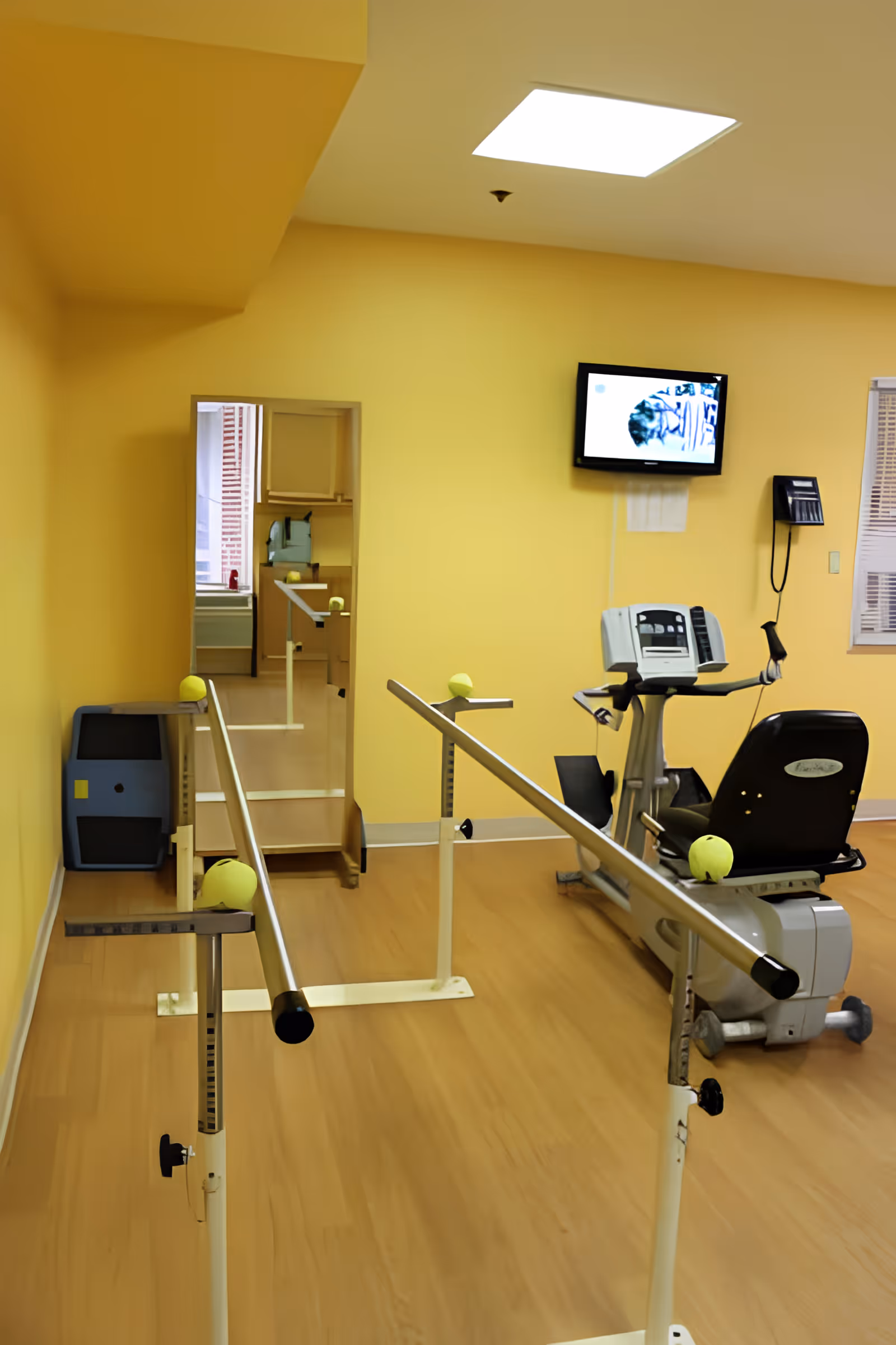 A bright yellow-walled therapy room with parallel walking bars, a recumbent exercise bike, a wall-mounted TV and a mirror.