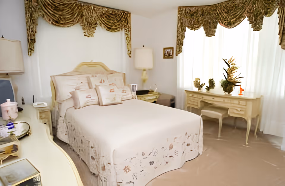 Bright bedroom with a double bed, matching vintage dressers and a vanity beneath large curtained windows.