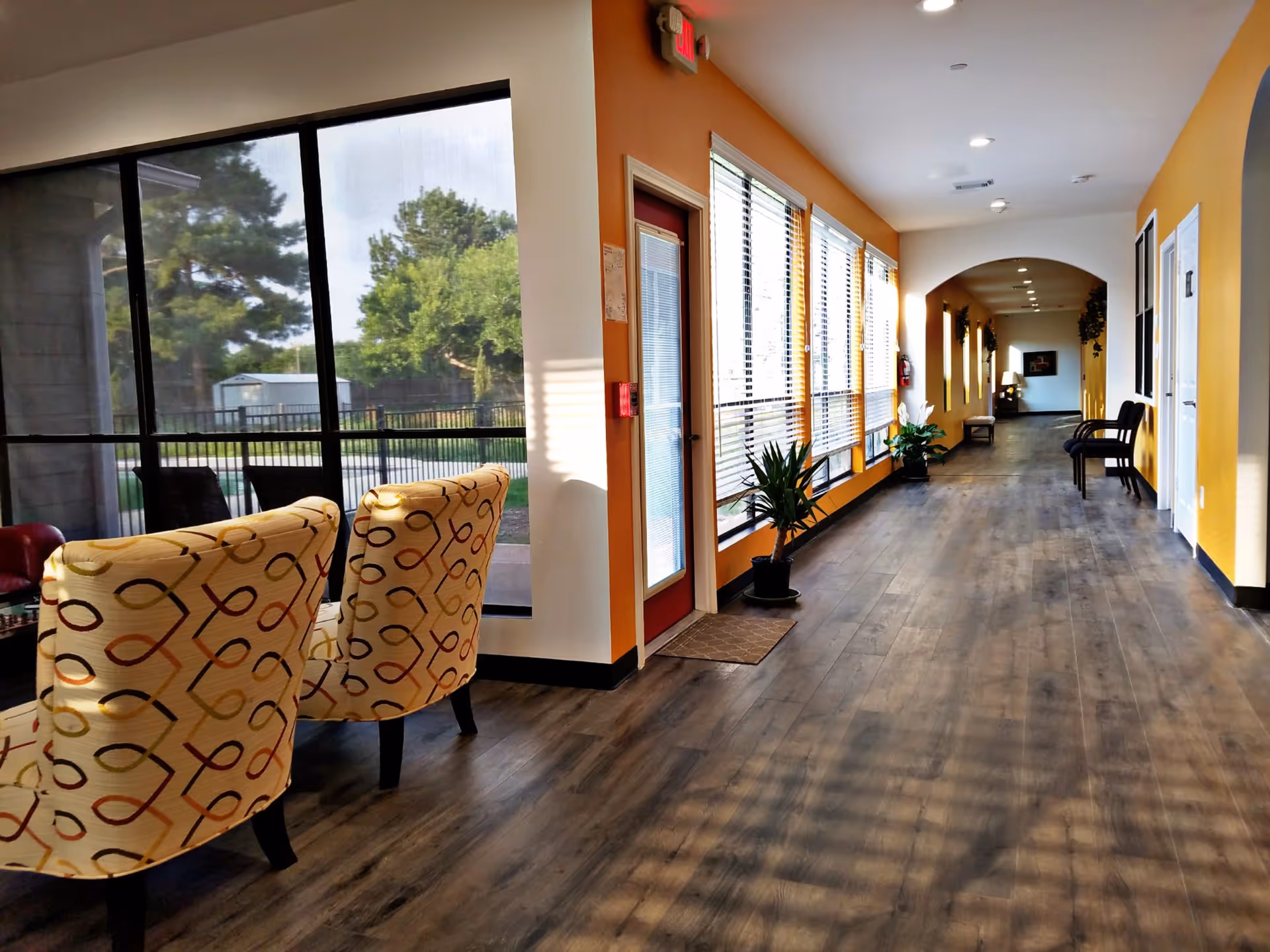 A bright hallway in a residential care facility with large windows on one side letting in natural light. The hallway has wood flooring, yellow and white walls, and several chairs and plants along the walls. Outside the windows, trees and a fenced area are visible.