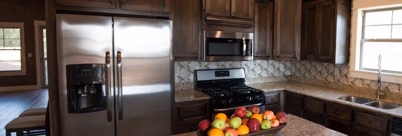 Modern kitchen with stainless steel refrigerator, microwave, and gas stove. The kitchen features dark wooden cabinets, granite countertops, a double sink under a window, and a bowl of assorted fresh fruits on the counter.