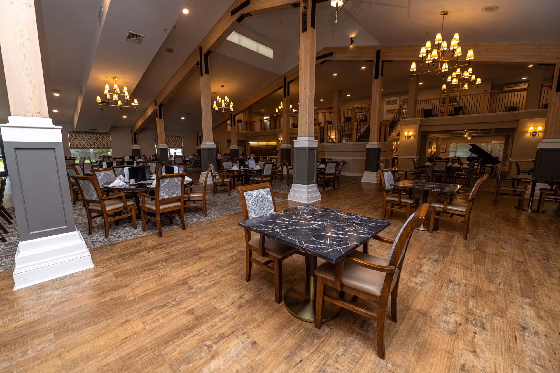Spacious dining room with multiple tables and chairs arranged neatly. The room features wooden floors, large wooden pillars, chandeliers, and a grand piano in the background. The area has a warm and inviting ambiance with soft lighting and a second-floor balcony overlooking the dining space.