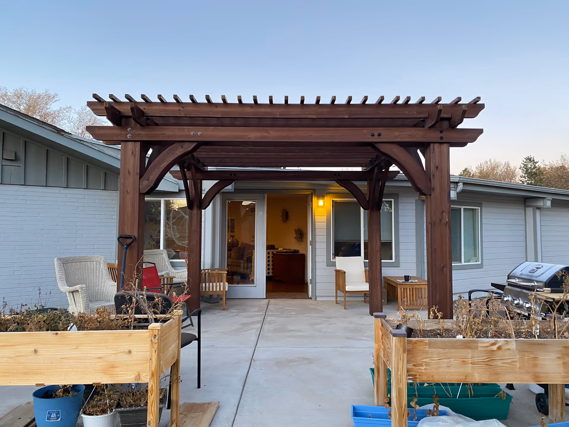 Outdoor patio area at Gardens Care Senior Living - Pinehurst featuring a wooden pergola with seating underneath, including wicker and wooden chairs. Raised garden beds with dried plants are on either side of the concrete patio, and a barbecue grill is visible on the right side. The building exterior is light gray with windows and a door leading inside, illuminated by a yellow porch light.