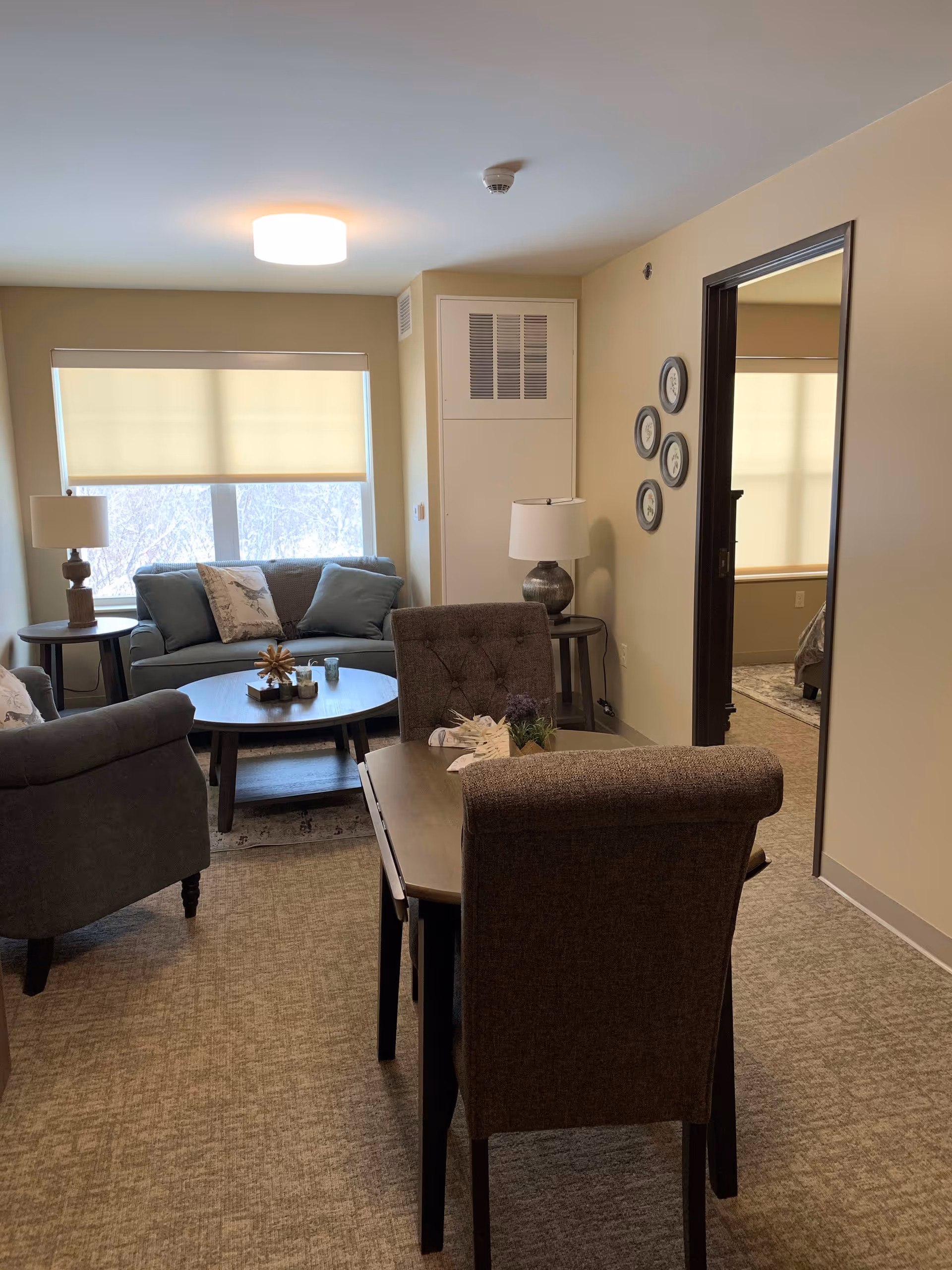 Small furnished living room in a senior apartment featuring a sofa, armchairs, a coffee table, dining table, and lamps.