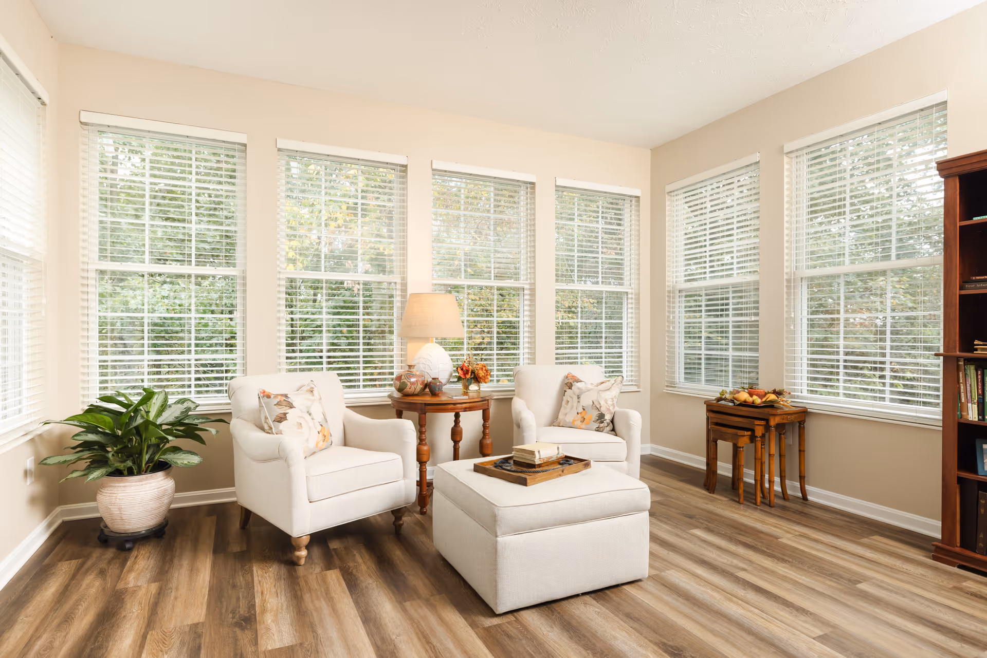 A bright and cozy living room area with large windows featuring white blinds. The room has two white upholstered armchairs with floral pillows, a matching ottoman with a tray on top, a wooden side table with a lamp and decorative items, a potted plant in a beige pot, and a wooden bookshelf filled with books. The floor is wood with a natural finish.