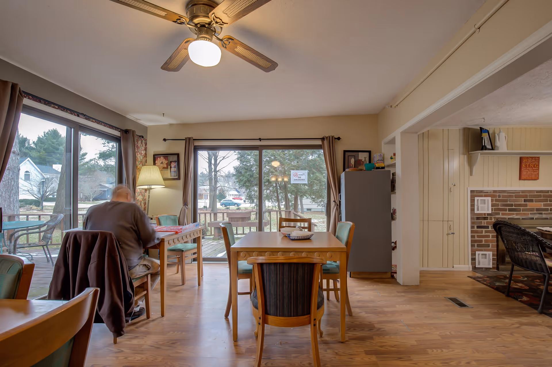 A cozy dining area in a senior living facility with wooden tables and chairs. A person is seated at one of the tables near large sliding glass doors that open to an outdoor deck. The room has a ceiling fan with a light, a floor lamp, framed artwork on the walls, and a refrigerator near a brick fireplace in an adjacent room.