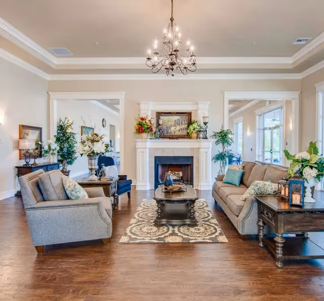A bright and spacious living room with a chandelier hanging from the ceiling. The room features a fireplace centered on the far wall with decorative plants and a framed painting above it. There are two upholstered sofas facing each other with patterned cushions, a dark wooden coffee table on a patterned rug, and side tables with lamps and floral arrangements. Large windows allow natural light to fill the room.