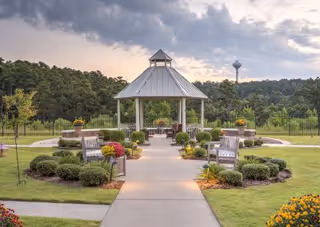 Concrete path leading to a white gazebo in a landscaped garden with benches, shrubs, and a distant water tower under a cloudy sky.