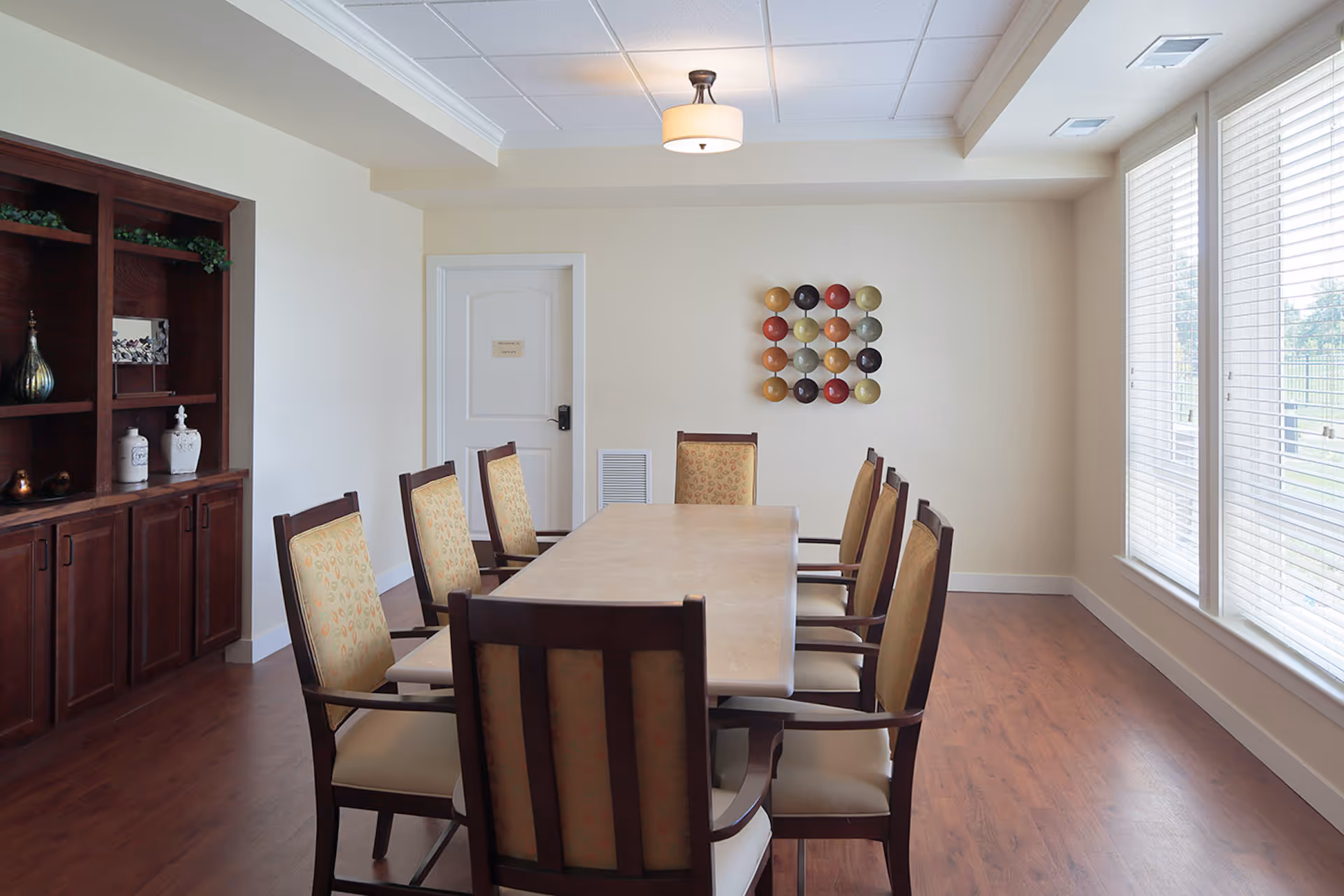 A bright dining room with a long rectangular table surrounded by eight cushioned chairs with wooden frames. The room has large windows with blinds on the right side, a wooden cabinet with decorative items on the left, and a colorful circular wall art piece on the far wall. The floor is wooden, and the ceiling has a light fixture.