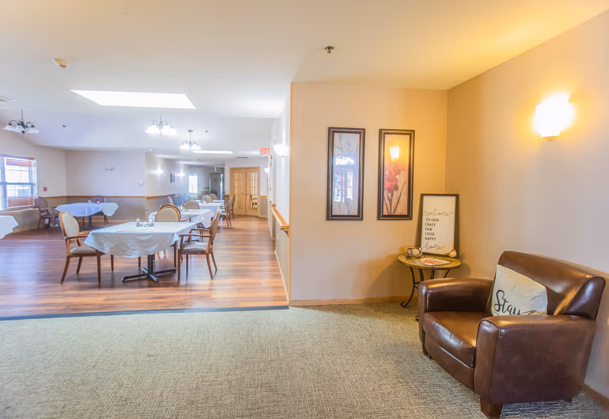 Interior view of a senior living facility showing a dining area with tables covered in white tablecloths and chairs arranged around them. To the right, there is a cozy corner with a brown leather armchair, a small round table with decorative items, and two framed floral artworks on the wall. The space is well-lit with ceiling lights and wall sconces.