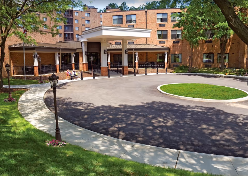Front exterior view of a multi-story brick building with a covered entrance and a circular driveway. There are trees and green grass surrounding the driveway, and a few elderly people sitting on benches near the entrance.