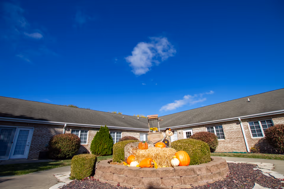 Courtyard with a raised circular planter decorated with pumpkins and a scarecrow in front of a single-story brick senior living building under a bright blue sky.