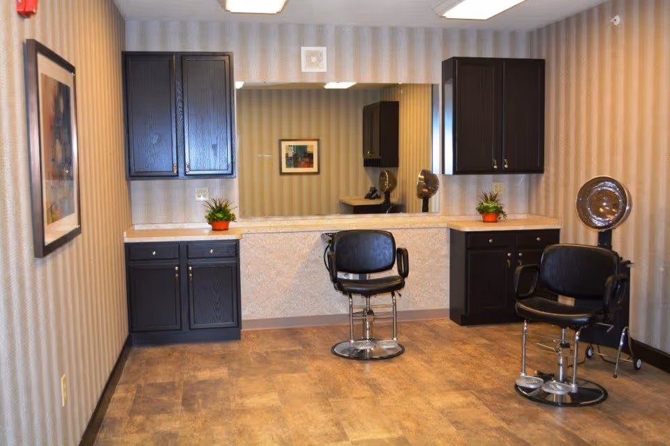 Interior view of a small salon area with two black salon chairs, a large mirror on the wall, dark wood cabinets, and a hair dryer hood. The room has beige striped wallpaper and a framed picture on the left wall.