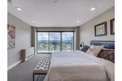 A spacious bedroom with a large bed featuring white bedding and multiple pillows. There is a cushioned bench at the foot of the bed. The room has light-colored walls, recessed ceiling lights, and a large sliding glass door that opens to a balcony with a scenic view of trees and hills in the distance. Two framed pictures hang on the wall above the bed.