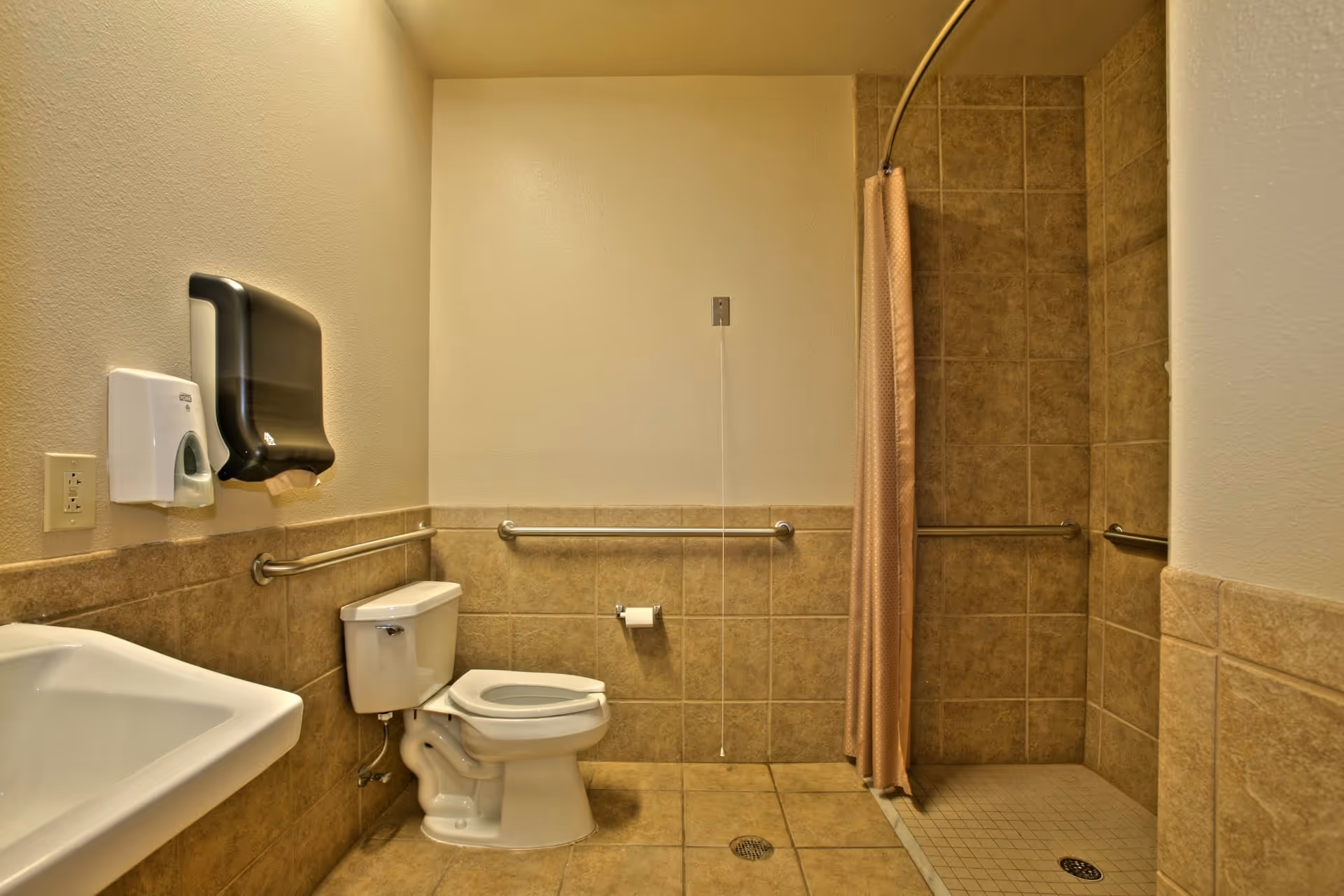 A clean, accessible bathroom with beige tiled walls and floor. The bathroom features a white toilet with grab bars on the walls around it, a white sink, a wall-mounted soap dispenser, a paper towel dispenser, and a walk-in shower with a beige curtain and grab bars inside.