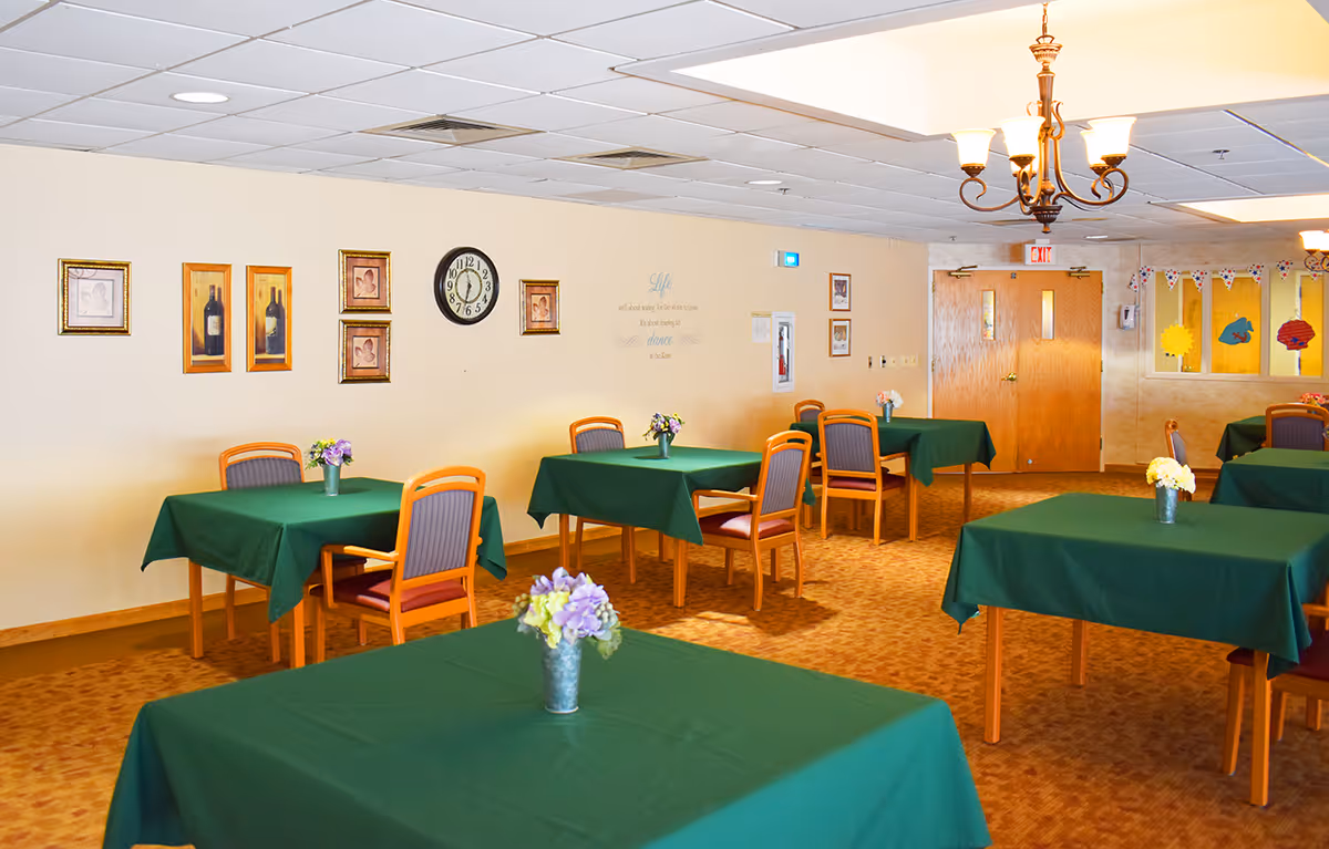 A bright dining room with multiple tables covered in green tablecloths, wooden chairs, floral centerpieces, wall art, and a chandelier.