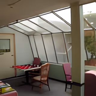 Indoor seating area with several chairs and a table near a large slanted window or skylight, allowing natural light to enter the room.
