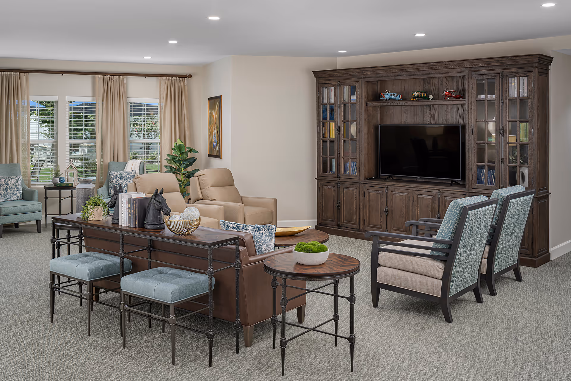 A spacious and well-lit living room area with a large wooden entertainment center housing a flat-screen TV. The room features comfortable seating including two beige recliners, a brown leather sofa with decorative pillows, two blue cushioned stools, and two armchairs with patterned upholstery. There are side tables with decorative items and a large window with beige curtains letting in natural light.