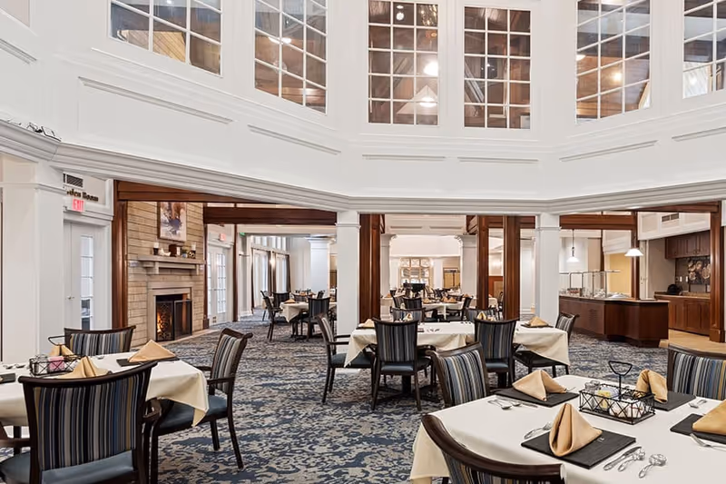 A spacious dining room with multiple tables covered in white tablecloths and beige napkins folded in a triangle shape. The room features a patterned carpet, wooden chairs with striped upholstery, a fireplace on the left side, and a buffet area on the right. Large windows near the ceiling allow natural light to fill the space.