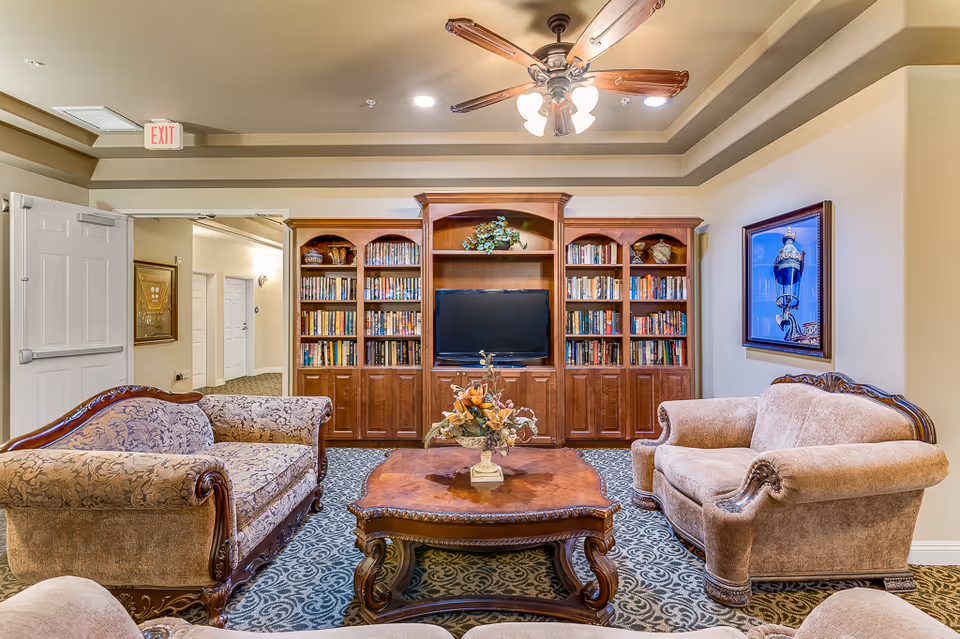 Cozy senior living common room with upholstered sofas, an ornate coffee table, built-in bookshelves and a TV under a ceiling fan.