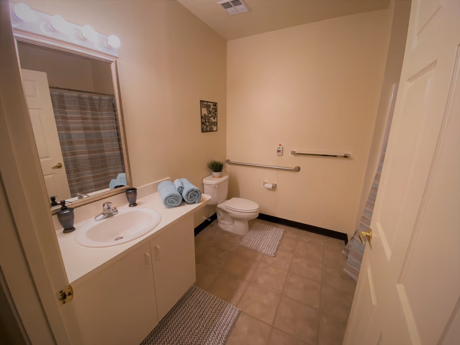 A clean bathroom with beige walls and tiled floor. There is a white sink with a countertop holding three rolled blue towels and two soap dispensers. Above the sink is a large mirror with lights. To the right is a toilet with a small plant on the tank and a grab bar on the wall. A striped shower curtain is partially visible behind the door.