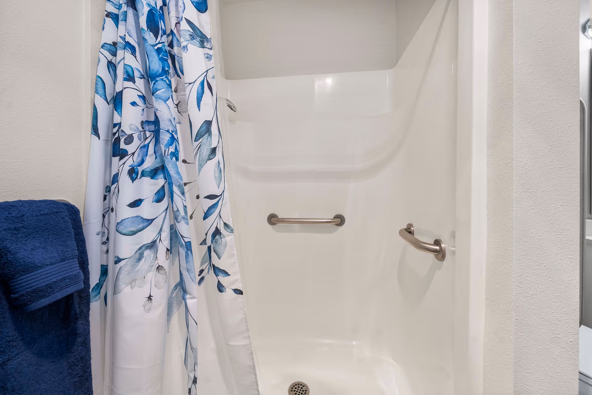 A white shower stall with two metal grab bars, one on the back wall and one on the side wall. A blue and white floral shower curtain is partially drawn to the left, and a dark blue towel hangs on a towel rack on the left wall.